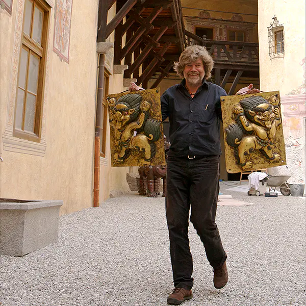Een man met krullend haar en een baard glimlacht terwijl hij over grind loopt, met in elke hand twee ingelijste kunstwerken met mythologische wezens, op een binnenplaats met sierlijke muren.